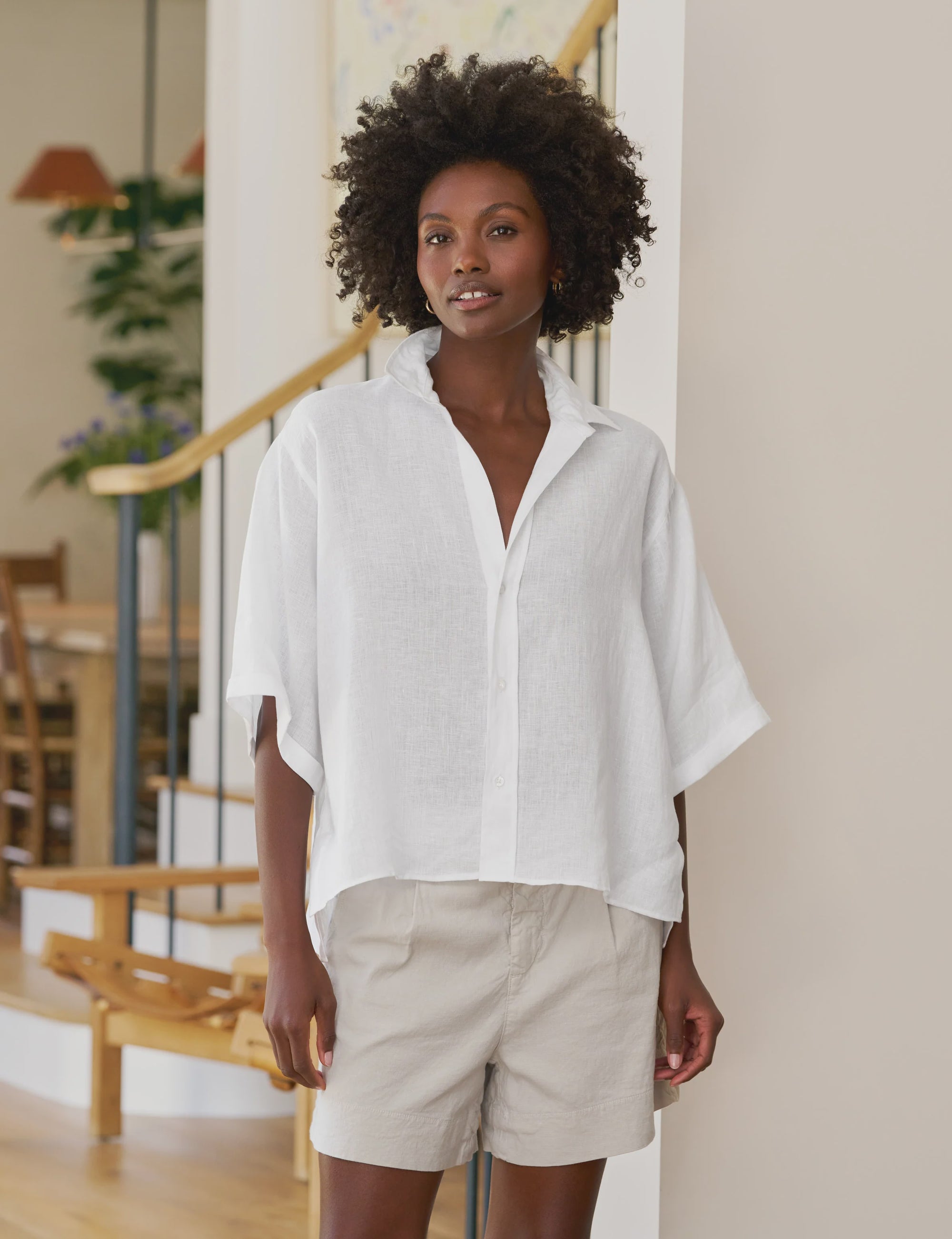 A woman with short curly hair wears the Frank & Eileen ROSIE SS Button Up F25 and light beige shorts, standing indoors in a modern, sunlit room with wooden stairs and plants in the background.