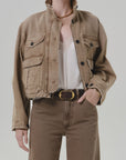 Tan Citizens of Humanity barn jacket with white lace top, brown pants, and gold-buckle belt in a modern setting.