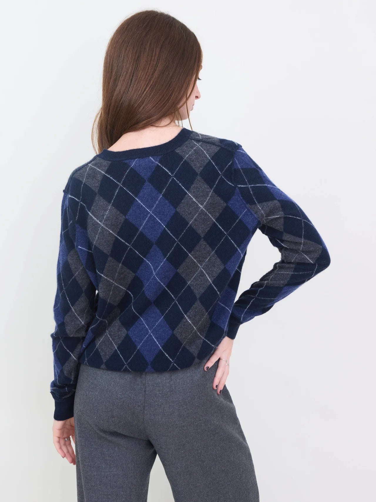 A person with long brown hair, seen from behind, wears the Brazeau Tricot Argyle V-Neck sweater in navy and gray with gray pants against a plain white background.