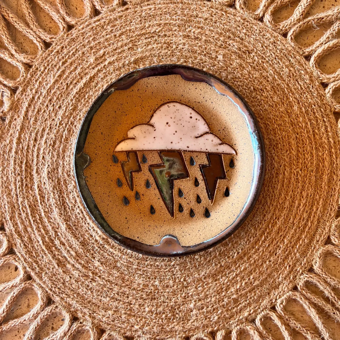 The Monsoon Trinket Dish by Faire features a round ceramic design with a cloud, raindrops, and metallic lightning bolt, displayed on a woven textured placemat—ideal for fans of unique handmade desert decor.