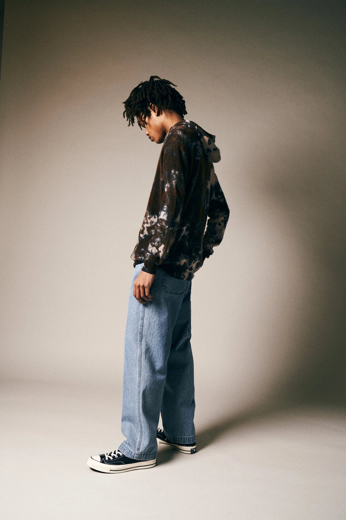 A person with short dreadlocks stands sideways, wearing a Riverside Tool & Dye Cashmere Hoodie, light blue wide-leg jeans, and black Converse sneakers against a neutral background.