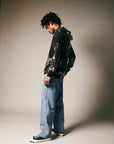 A person with short dreadlocks stands sideways, wearing a Riverside Tool & Dye Cashmere Hoodie, light blue wide-leg jeans, and black Converse sneakers against a neutral background.