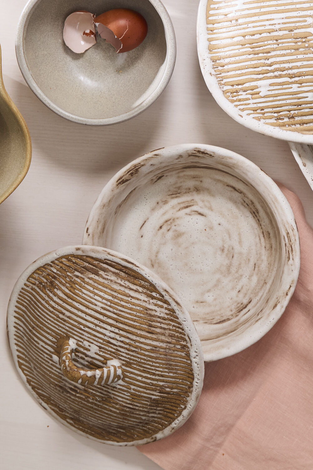 A round Accent Decor Sycamore Lidded Dish with a bark-textured lid sits on a pink cloth, accompanied by a bowl of brown eggshells and an earthy ceramic dish on a light surface.