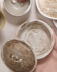 A round Accent Decor Sycamore Lidded Dish with a bark-textured lid sits on a pink cloth, accompanied by a bowl of brown eggshells and an earthy ceramic dish on a light surface.