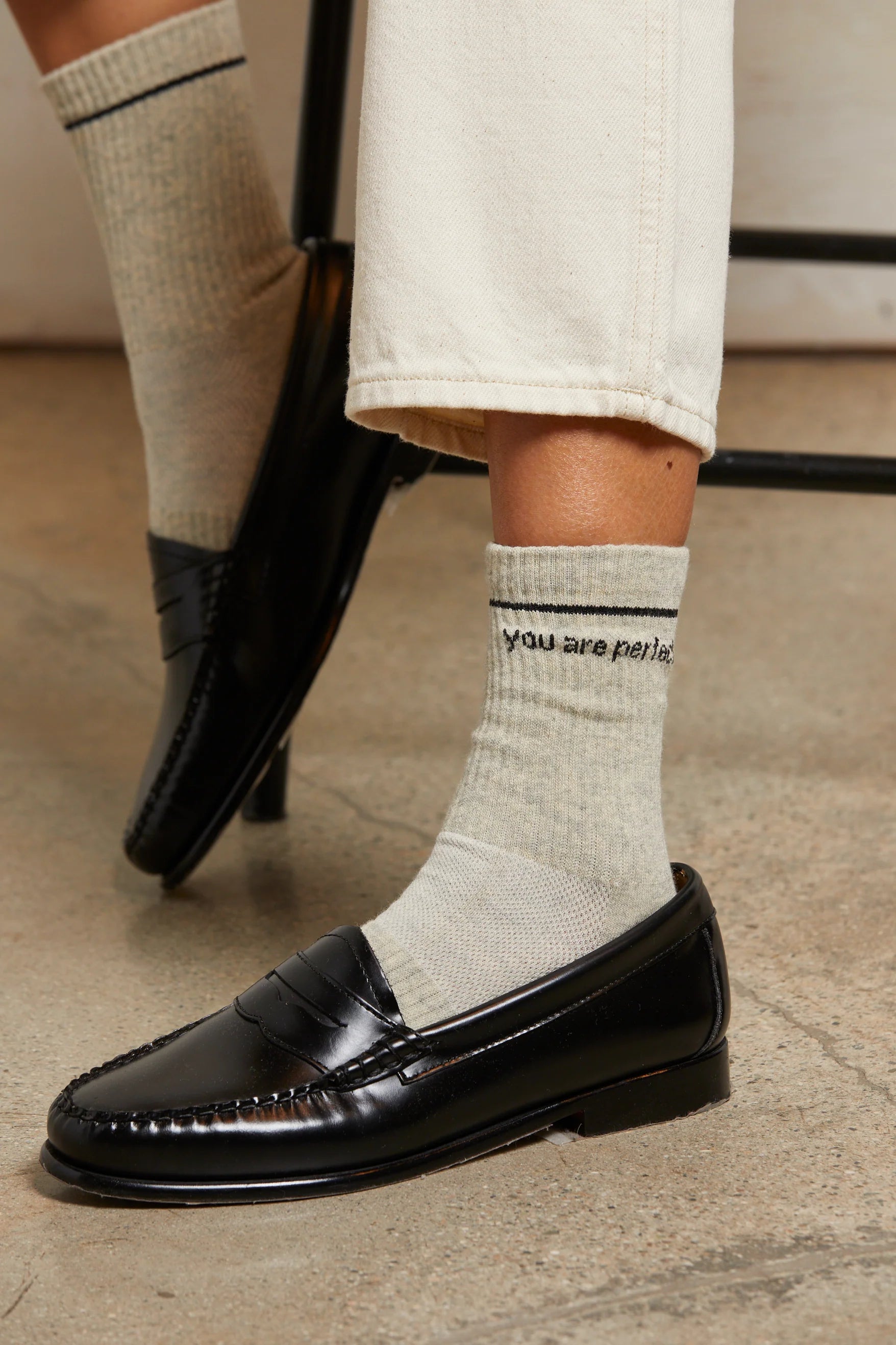 Someone wearing cream pants, black loafers, and Perfectwhitetee's You Are Perfect Sock—a vintage-inspired light gray ribbed cotton style with "you are perfect." in black text—sits cross-legged, showing off the visible wording on their sock.