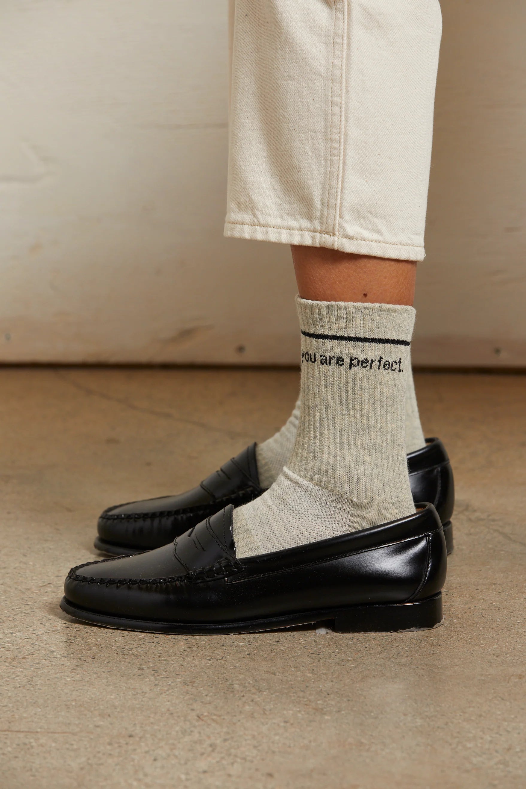 A person stands on a concrete floor against a light wall, wearing light beige pants, Perfectwhitetee's You Are Perfect Sock (vintage-inspired ribbed cotton socks with "you are perfect"), and black loafers.