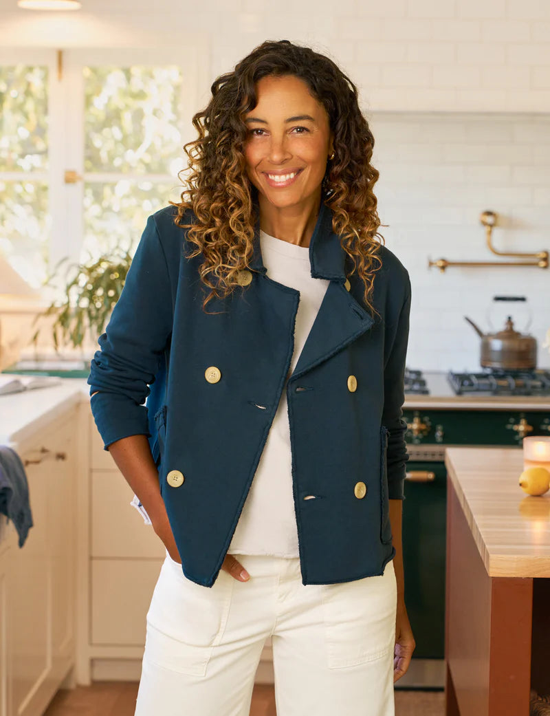 A woman with curly hair, wearing the Frank & Eileen MINI BELFAST Crop Peacoat in navy and white pants, smiles in a bright, modern kitchen with white cabinets and a green stove in the background.