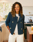 A woman with curly hair, wearing the Frank & Eileen MINI BELFAST Crop Peacoat in navy and white pants, smiles in a bright, modern kitchen with white cabinets and a green stove in the background.