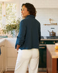 A woman with curly hair stands in a bright kitchen with white tiles and a green stove, wearing the Frank & Eileen MINI BELFAST Crop Peacoat and white pants, her hands in her pockets as she faces away.