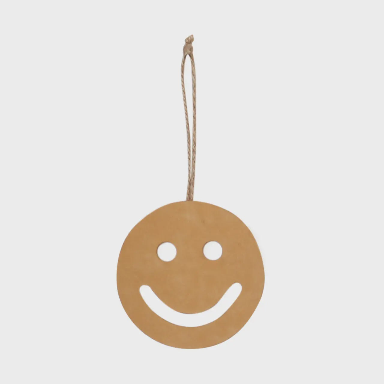 The Smiley leather ornament by Faire, crafted from repurposed leather and featuring a tan-colored smiley face with cut-out eyes and mouth, is suspended by a thin brown cord against a plain white background.