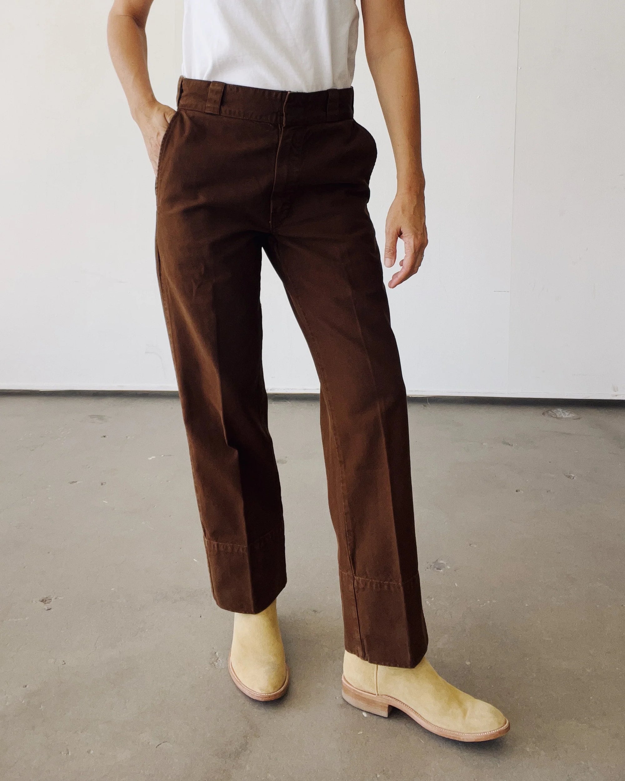 A person stands indoors on a concrete floor wearing Hey Gang's The Work Pant in brown cotton canvas, paired with a white shirt and light tan ankle boots. Their left hand is in their pocket, and their head is out of the frame.