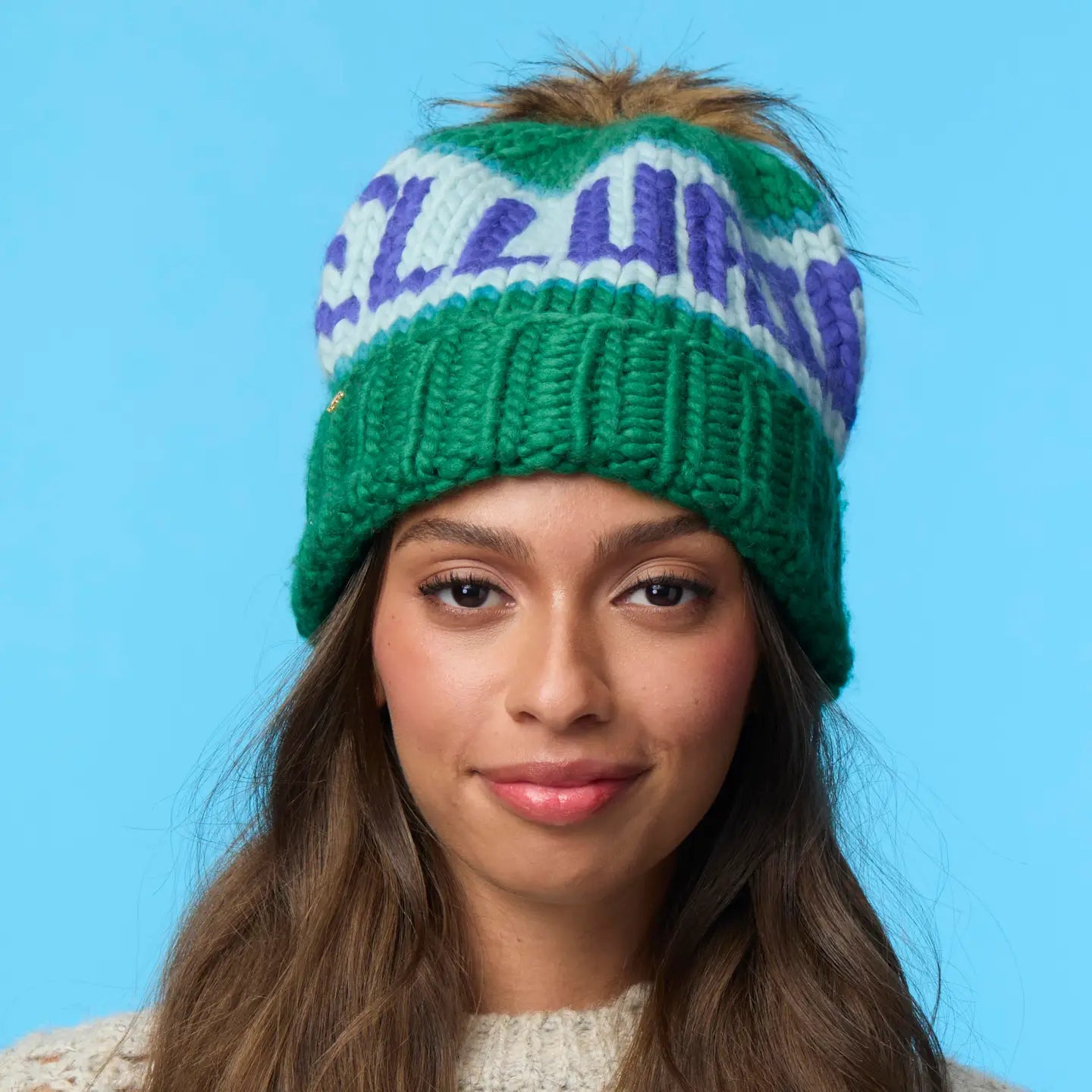 A woman with long brown hair wears the Faire Knit Ski Hat, a merino wool beanie with a faux fur pom pom, and a cream sweater against a light blue background.