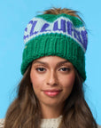 A woman with long brown hair wears the Faire Knit Ski Hat, a merino wool beanie with a faux fur pom pom, and a cream sweater against a light blue background.
