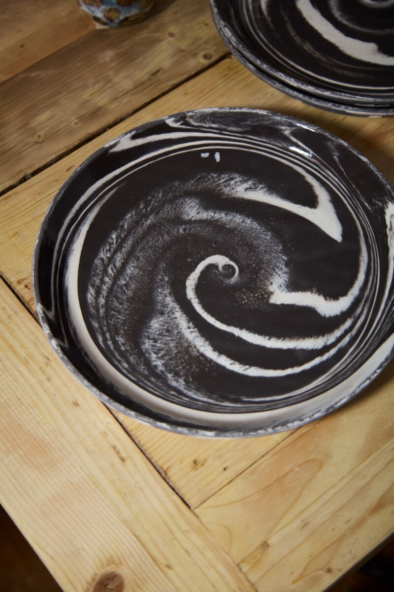 The Marbled Charger by Accent Decor, featuring a handcrafted black stoneware surface with swirling white patterns, sits on a wooden table. Another decorative platter is partially visible in the background.