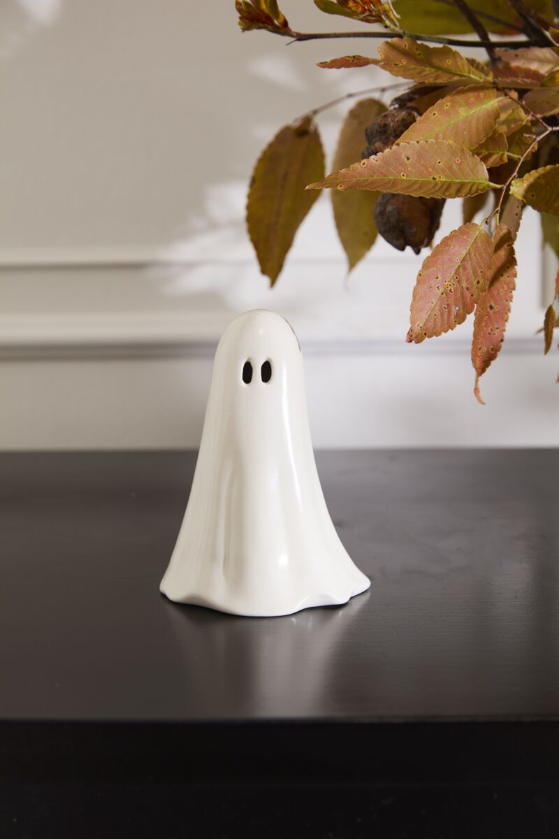 The Accent Decor ghostly Figurine Small is a hand-painted, white ceramic ghost with black eyes, perfect for charming Halloween décor. It sits on a dark surface with autumn leaves hanging in the top right corner of the image.