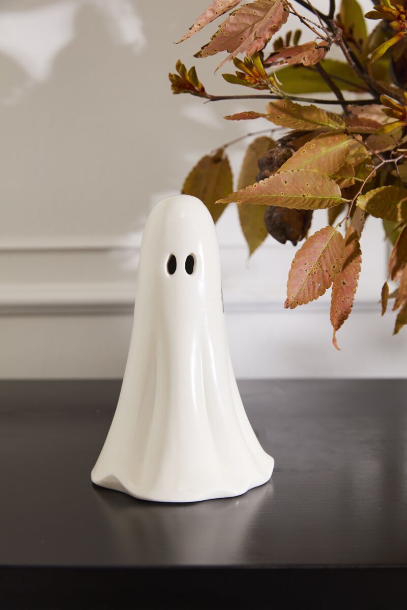 The Accent Decor Ghostly Figurine Large, a white ceramic ghost with black eye holes, is displayed on a black surface beside autumn leaves in brown, orange, and yellow—ideal for Halloween décor.