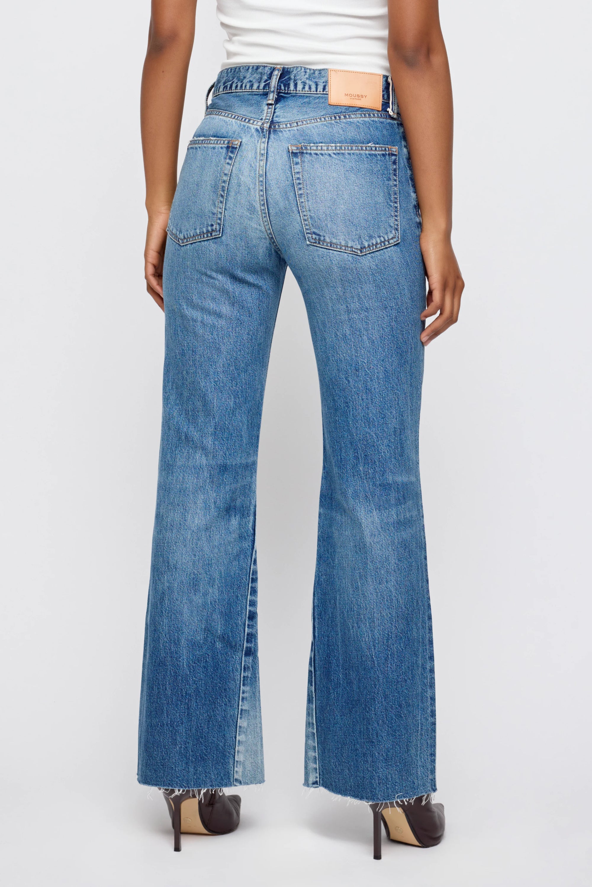 Someone is seen from behind wearing Moussy Asheville Remake Flare high-waisted blue jeans with frayed hems and front slits, paired with a white top and brown-heeled shoes, standing against a plain light background.