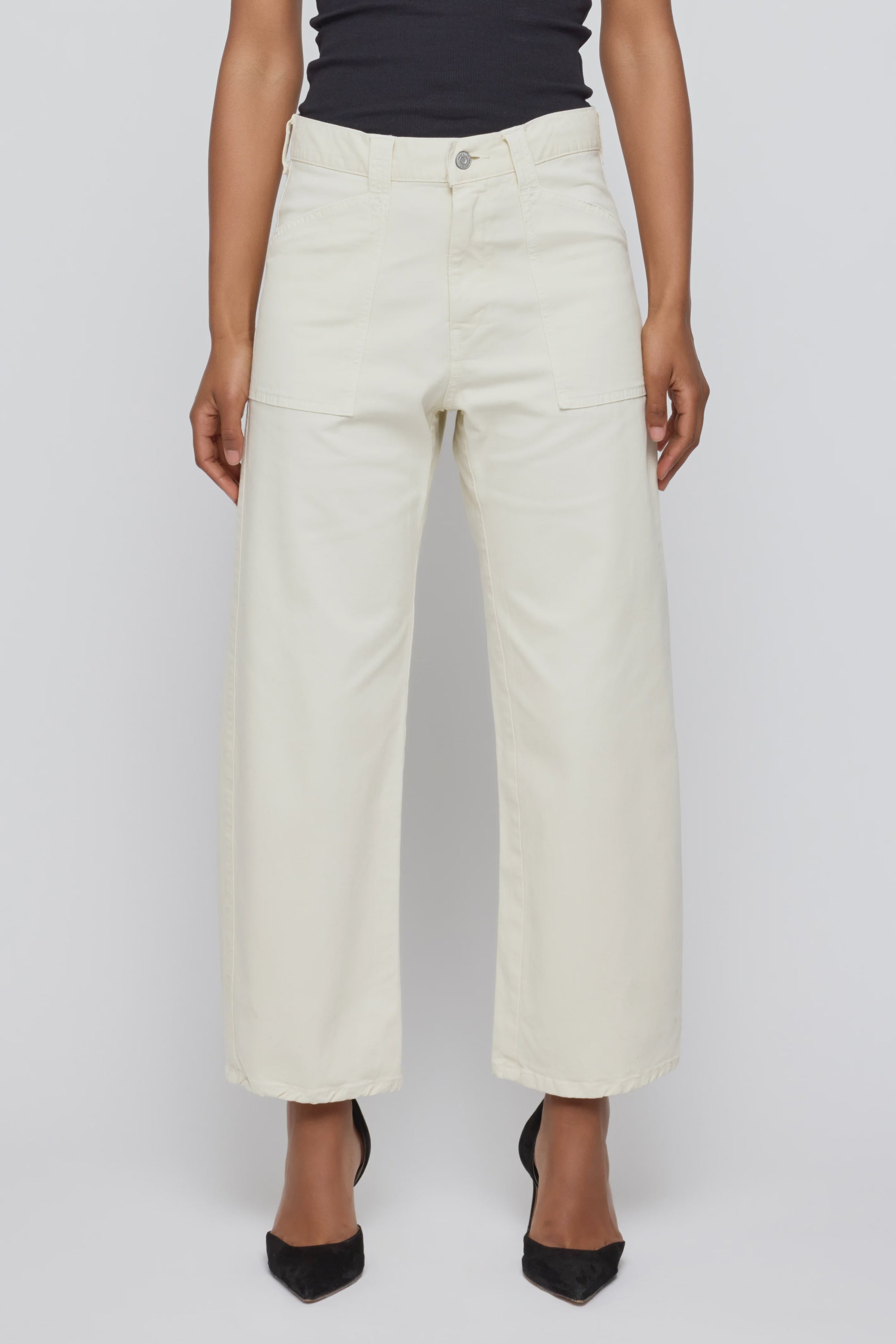 A person stands against a plain light background wearing Moussy Pomonok Gusset Cargo jeans in off-white with front patch pockets, paired with a black top and black pointed-toe heels.