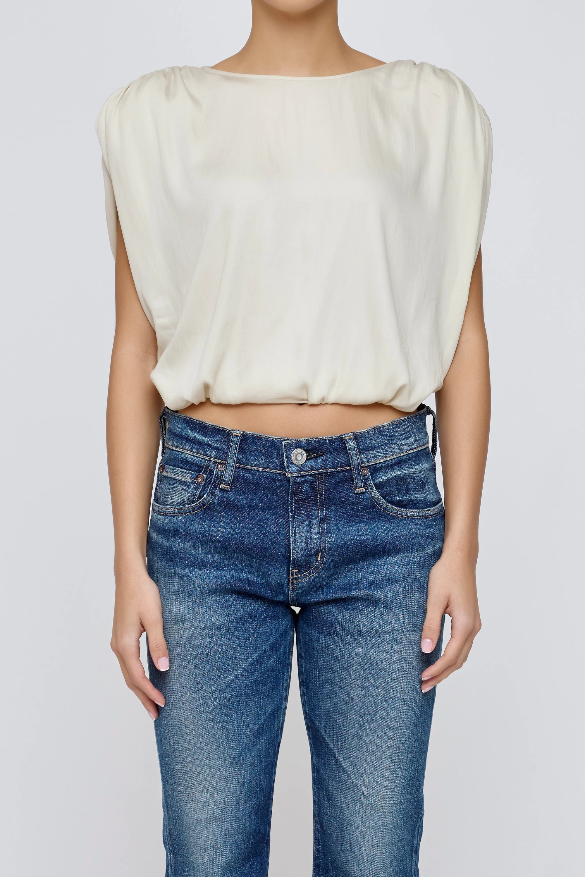 A person faces forward with arms relaxed at their sides, wearing the Moussy MV Satin Shell Cap Top in off-white and blue jeans. The background is plain light gray.