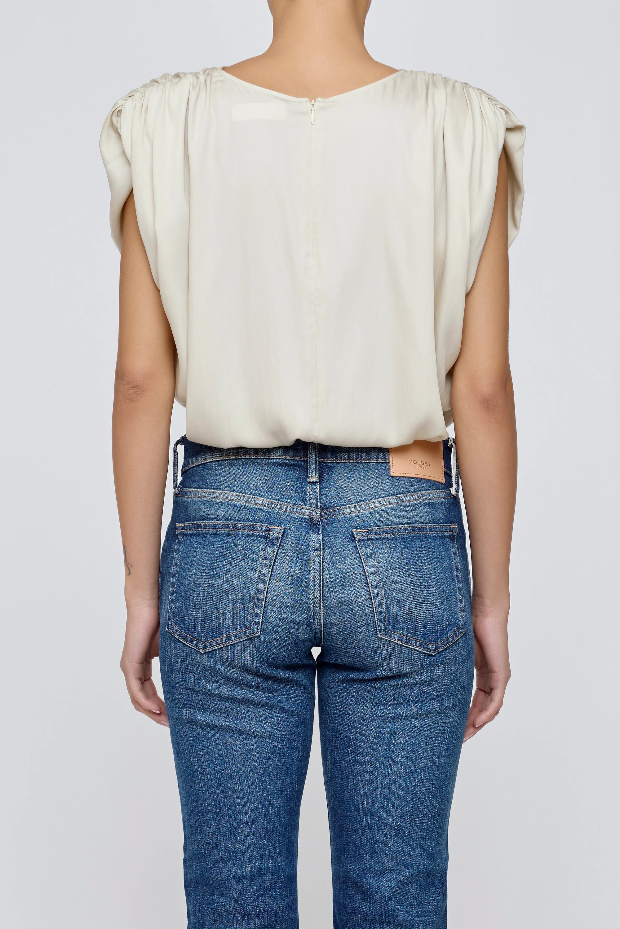 A person is seen from behind wearing the Moussy MV Satin Shell Cap Top—a light-colored, short-sleeved blouse—paired with blue jeans against a plain, light gray background.