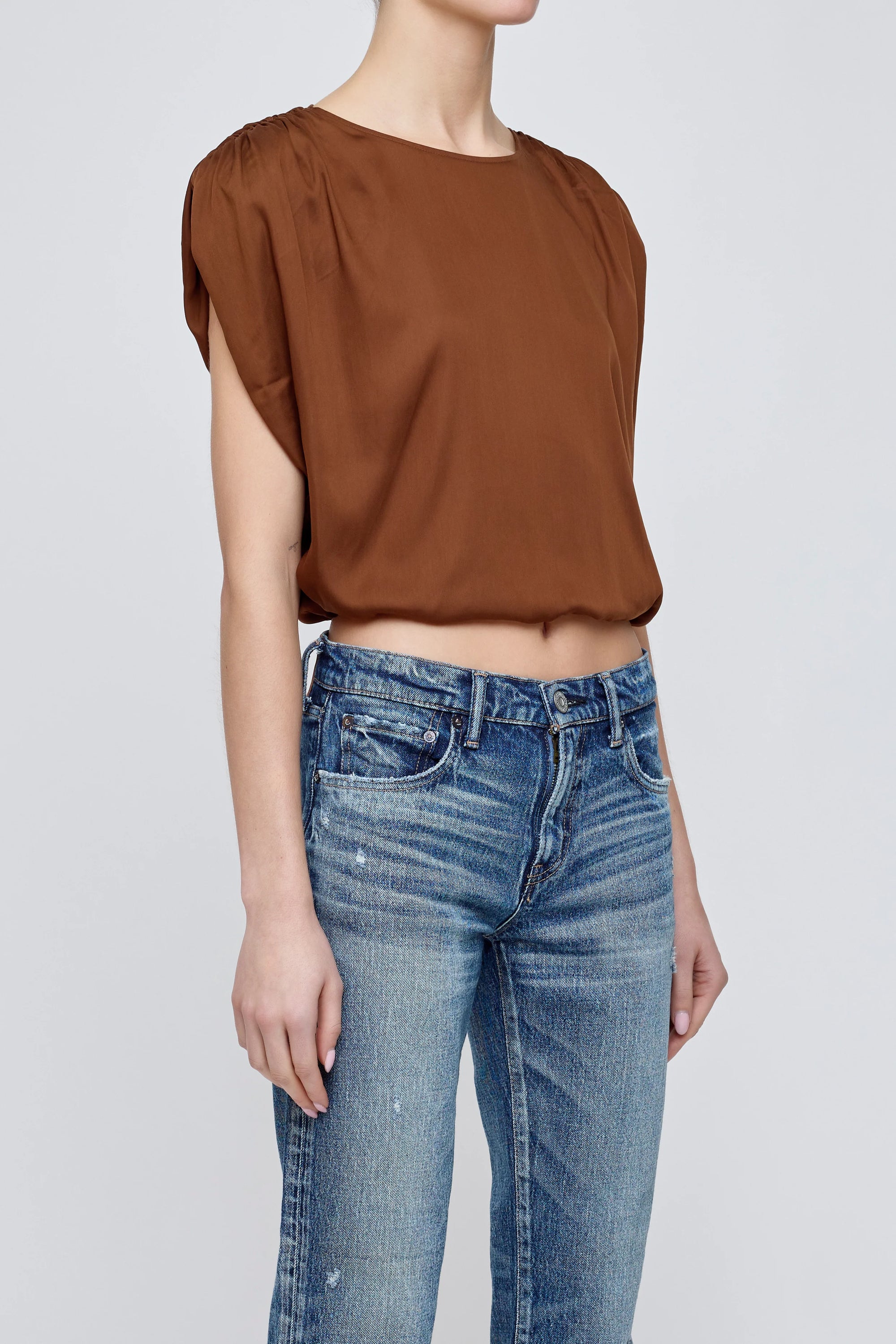 A person wearing the Moussy MV Satin Shell Cap Top—a loose brown, versatile short-sleeve piece—with blue jeans stands against a plain light gray background. The face is not visible.
