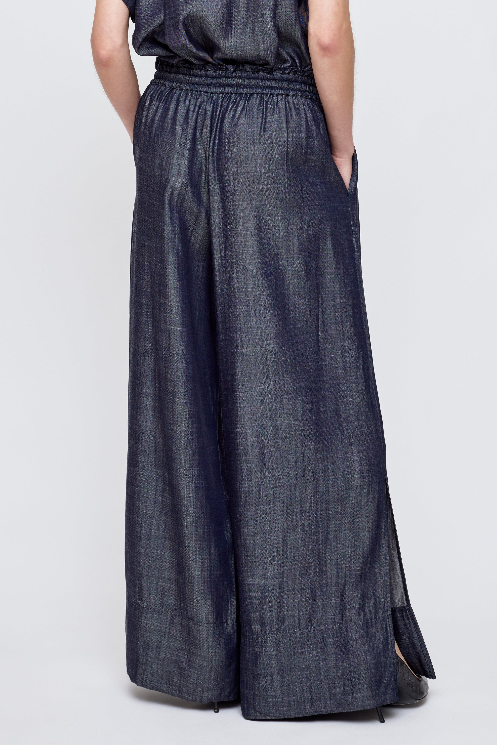 Person wearing Moussy High Waist Denim with wide legs, elastic waistband, side hem slits, pockets, and a subtle sheen, standing with hands in pockets. Photo shows the back view.