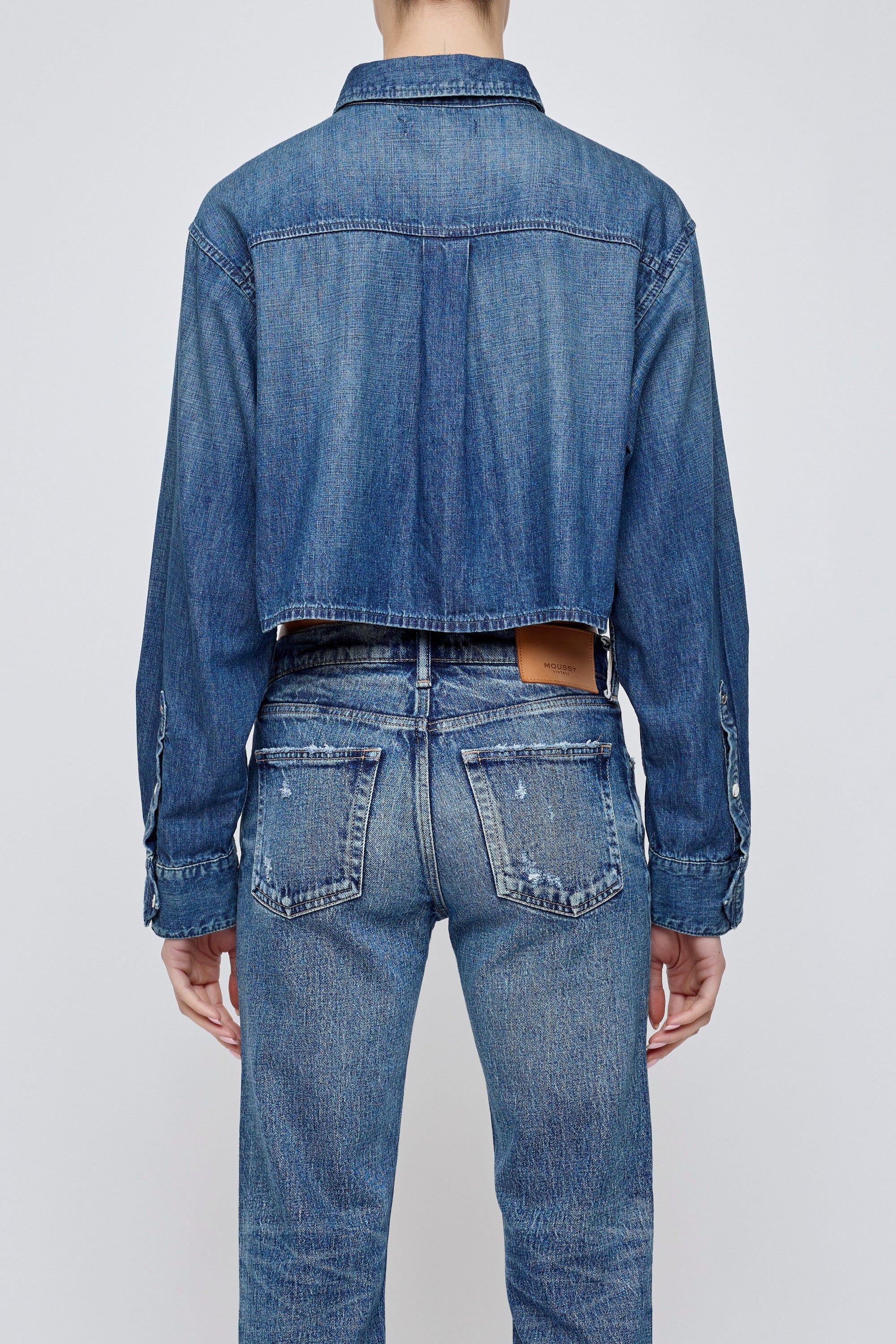 A person wearing the Moussy Foley Shirt, a cropped denim button-down, with blue jeans, viewed from the back against a plain light background.