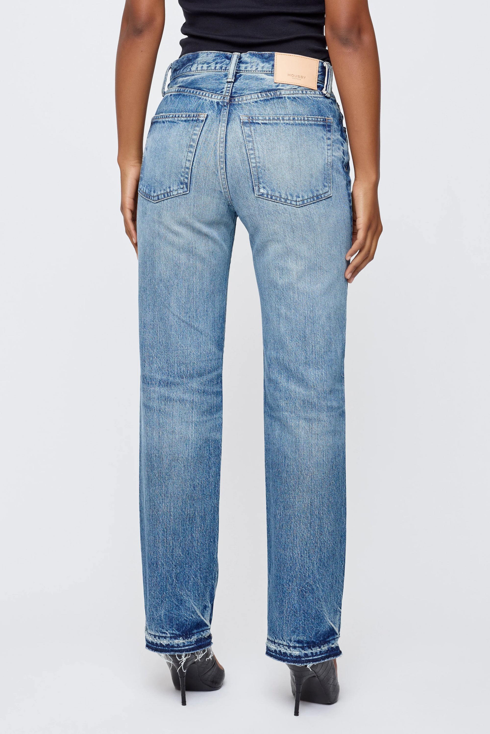 A person is shown from the back wearing Moussy’s Dorchester Straight mid-rise, full-length jeans with a black top and black pointed-toe shoes, standing with arms relaxed at their sides.