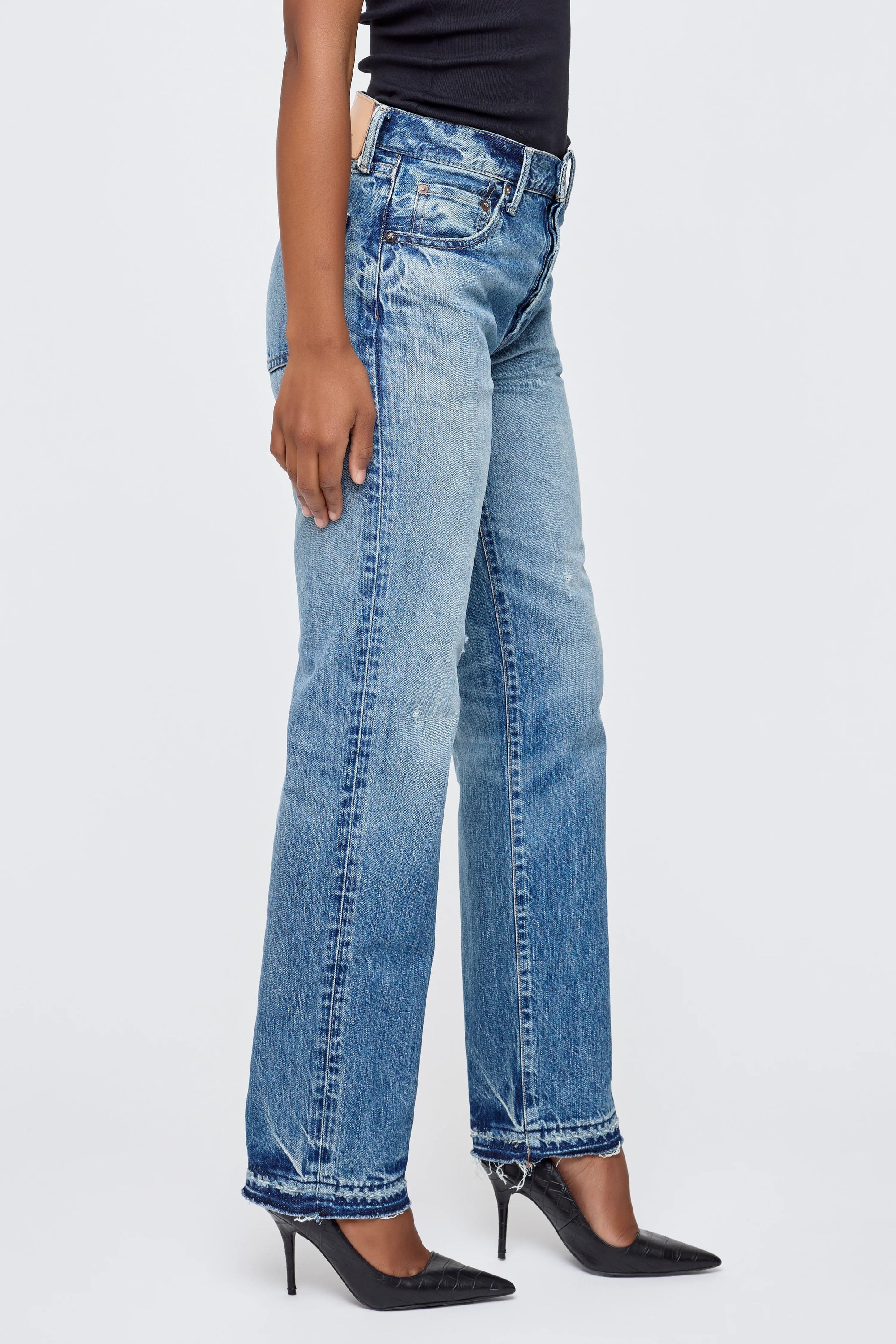 A person wearing the Moussy Dorchester Straight light blue mid rise jeans with black pointed-toe heels stands against a plain light background, showing only the lower half of their body.