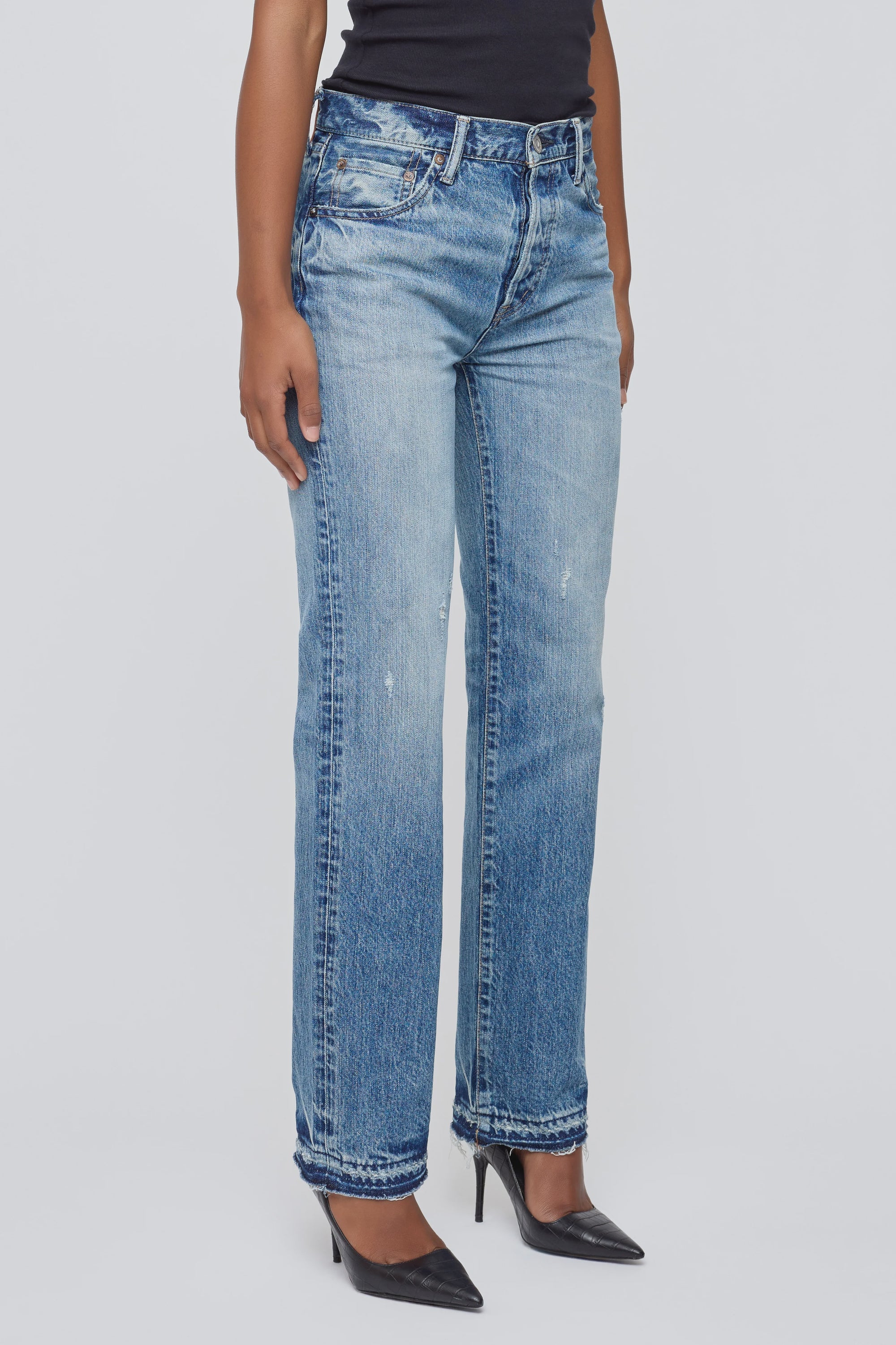 A person wears Moussy’s Dorchester Straight mid-rise jeans in light blue 100% cotton with black pointed-toe heels, posing against a plain white background. Only the lower body is shown.