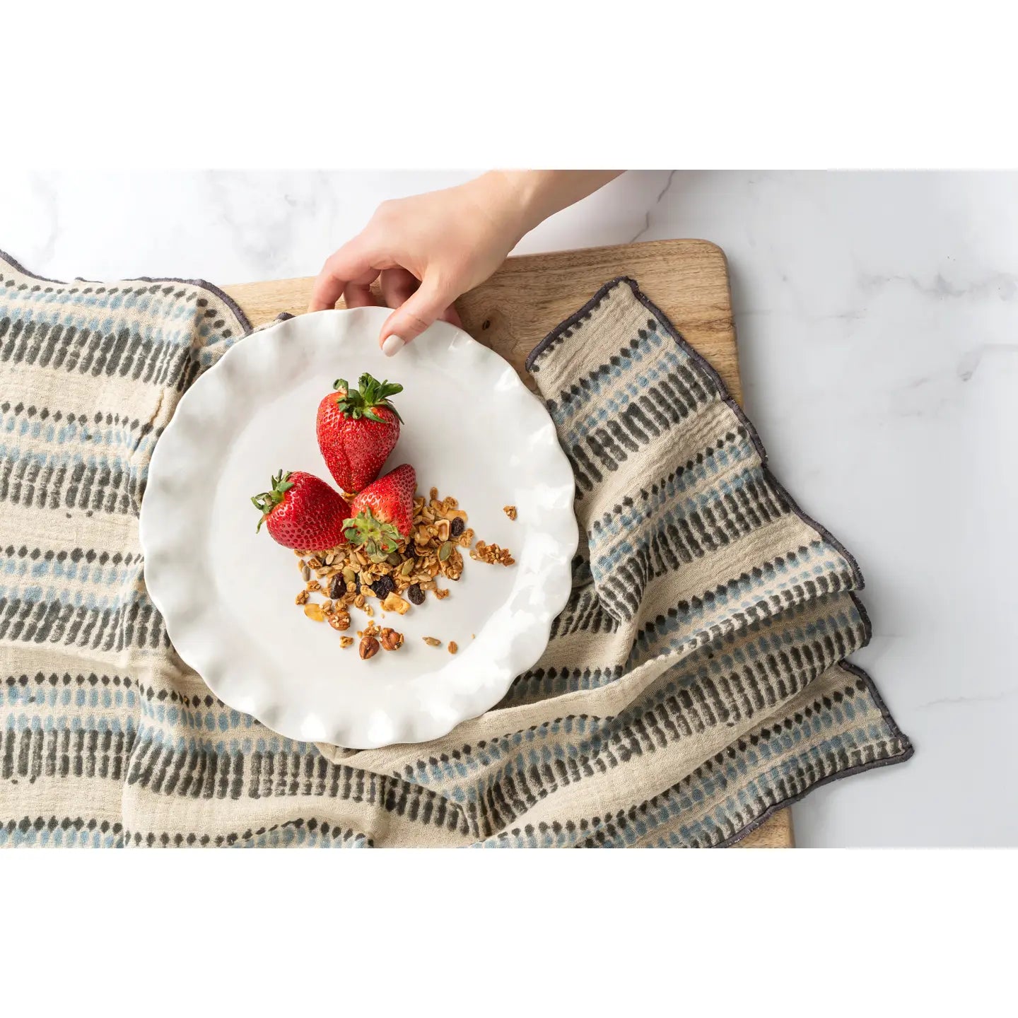 A hand sets a white plate with three strawberries and granola on the Ishita Block Print Tea Towel by Indaba, which features stripes and rests on a wooden surface.