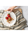 A hand sets a white plate with three strawberries and granola on the Ishita Block Print Tea Towel by Indaba, which features stripes and rests on a wooden surface.
