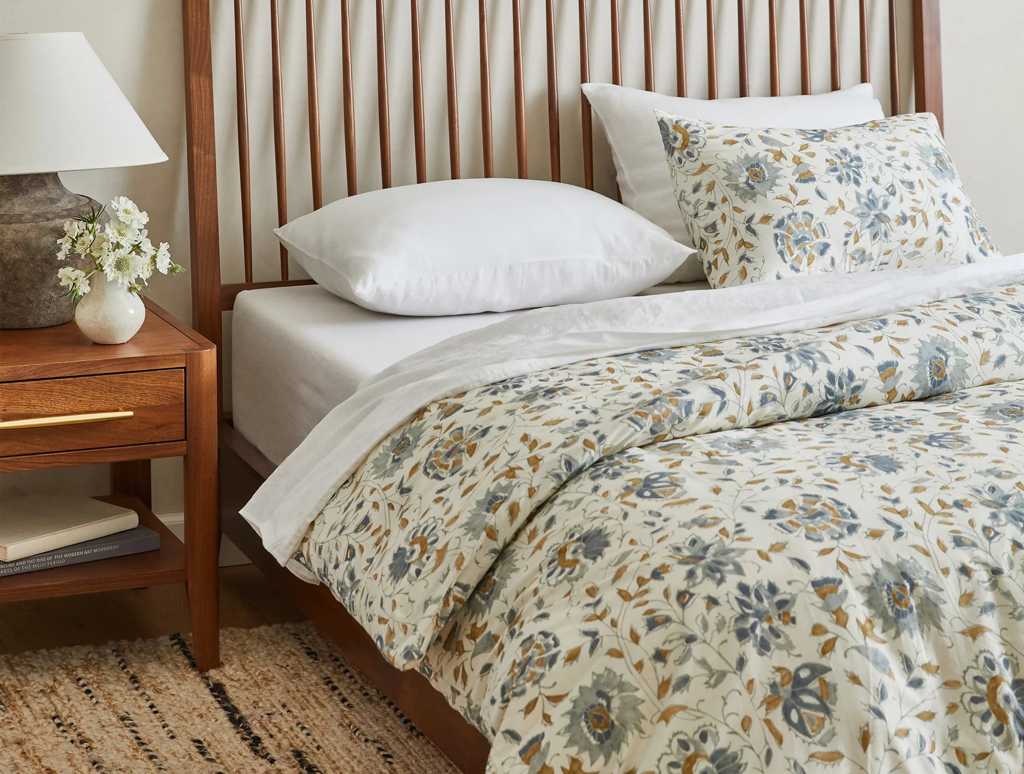 A bed is styled with Coyuchi Inc's Flora Org Print Duvet Undyed Lagoon King/CKing, showcasing a blue and yellow botanical design, beside a wooden nightstand holding a lamp, flowers in a vase, and a book on top of a woven rug.