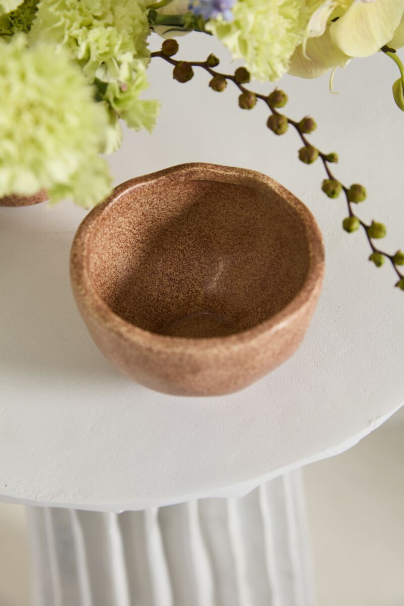 The Cyrus Bowl by Accent Decor, a small round terracotta piece, rests on a white textured surface with green and yellow flowers and leafy stems partially visible around it.