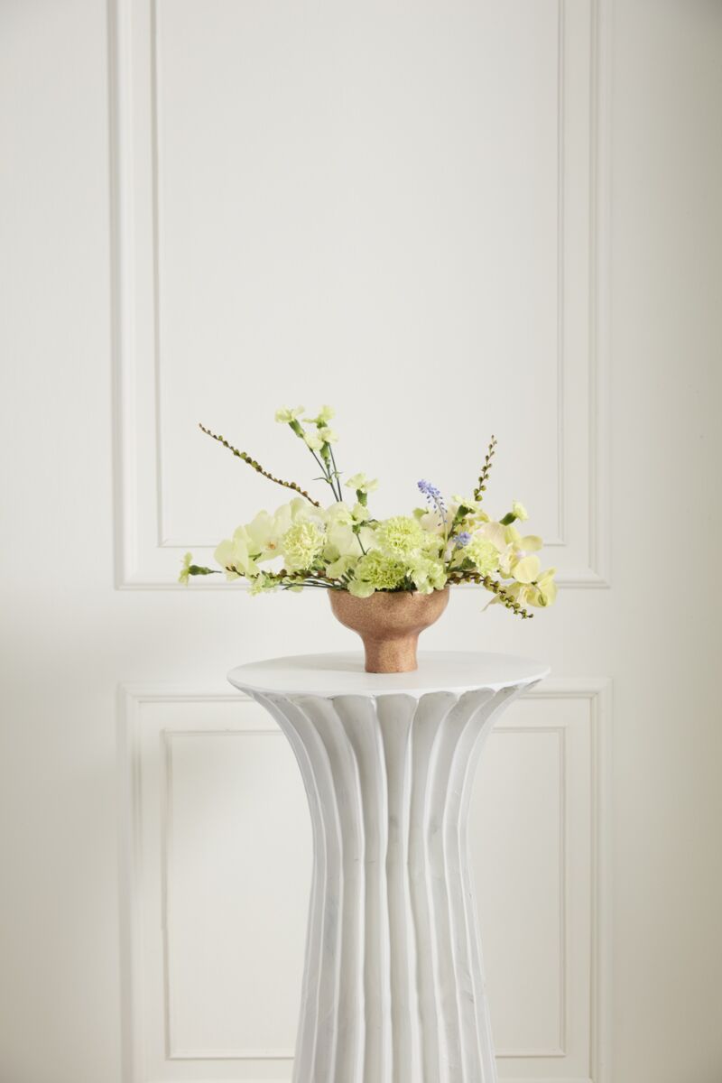 A minimalist arrangement of pale yellow and white blooms in the Cyrus Compote by Accent Decor sits on a white fluted pedestal against a white paneled wall.
