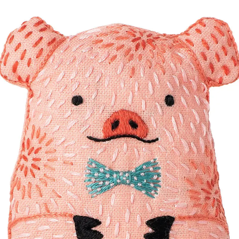 The Faire Pig Embroidery Kit includes pink fabric, black stitched eyes, a detailed snout, and a teal bow tie—ideal for beginner embroidery fans or anyone looking to create a plush pig toy.