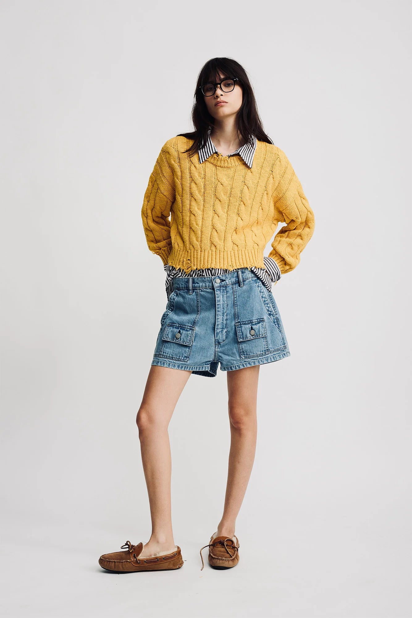 A woman with straight dark hair and glasses poses against a plain background in a Denimist Boxy Cable Sweater, layered over a striped shirt, denim shorts with large front pockets, and brown boat shoes.