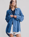 A woman wears the Denimist SUTTON BUTTON FRONT BRIGHT BLUE shirt over a black top and white frayed shorts, standing against a light background with her hands touching her shirt, looking slightly to the side.