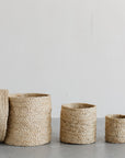 Three Faire Round Lidded Jute Baskets in different sizes are lined up on a gray surface with a plain white background. The largest basket's lid is propped against it.