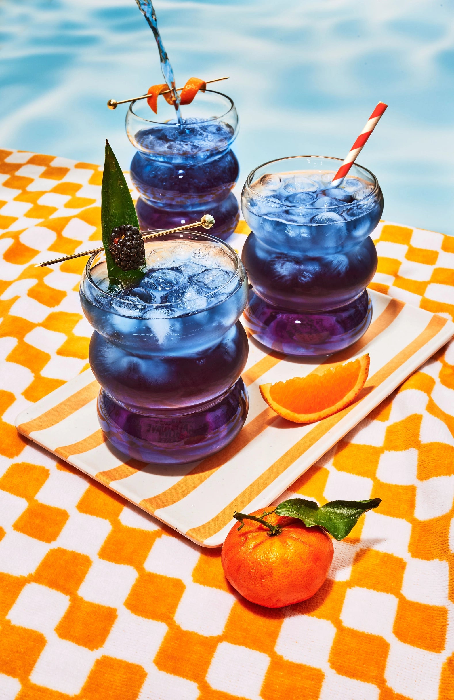 Three Faire Blackberry Jasmine Iced Teas sit in round glasses on a striped tray by a pool, garnished with fruit and striped straws. An orange slice and tangerine rest on an orange-and-white towel nearby.