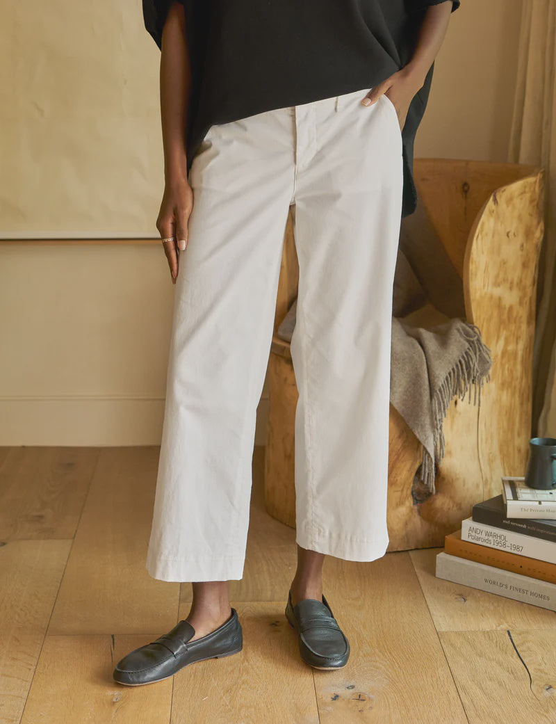 A person in white, mid-rise Frank & Eileen Westport Italian wide-leg pants, a black top, and black loafers stands on a wooden floor. A wooden chair with a folded blanket and stacked books is in the background.