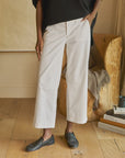 A person in white, mid-rise Frank & Eileen Westport Italian wide-leg pants, a black top, and black loafers stands on a wooden floor. A wooden chair with a folded blanket and stacked books is in the background.