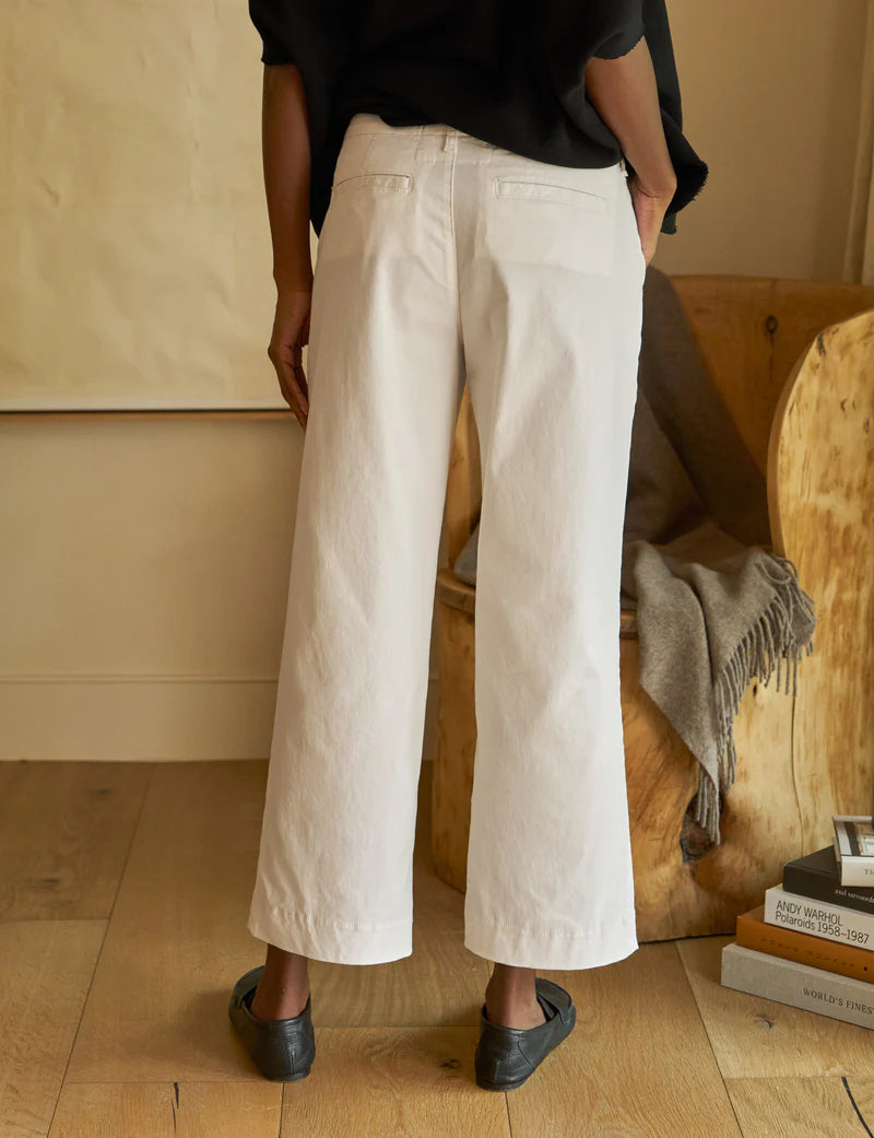 A person stands with their back to the camera, wearing Frank & Eileen's Westport Italian wide-leg pants and black shoes. They are in a room with wooden flooring, next to a wooden chair draped with a gray blanket and a small stack of books.