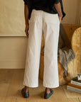 A person stands with their back to the camera, wearing Frank & Eileen's Westport Italian wide-leg pants and black shoes. They are in a room with wooden flooring, next to a wooden chair draped with a gray blanket and a small stack of books.