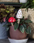A potted plant displays the Accent Decor White Spore Plant Stick with a ceramic toadstool design—one white with brown spots, one red with white—amid green leaves, while woven baskets and another plant decorate the background.