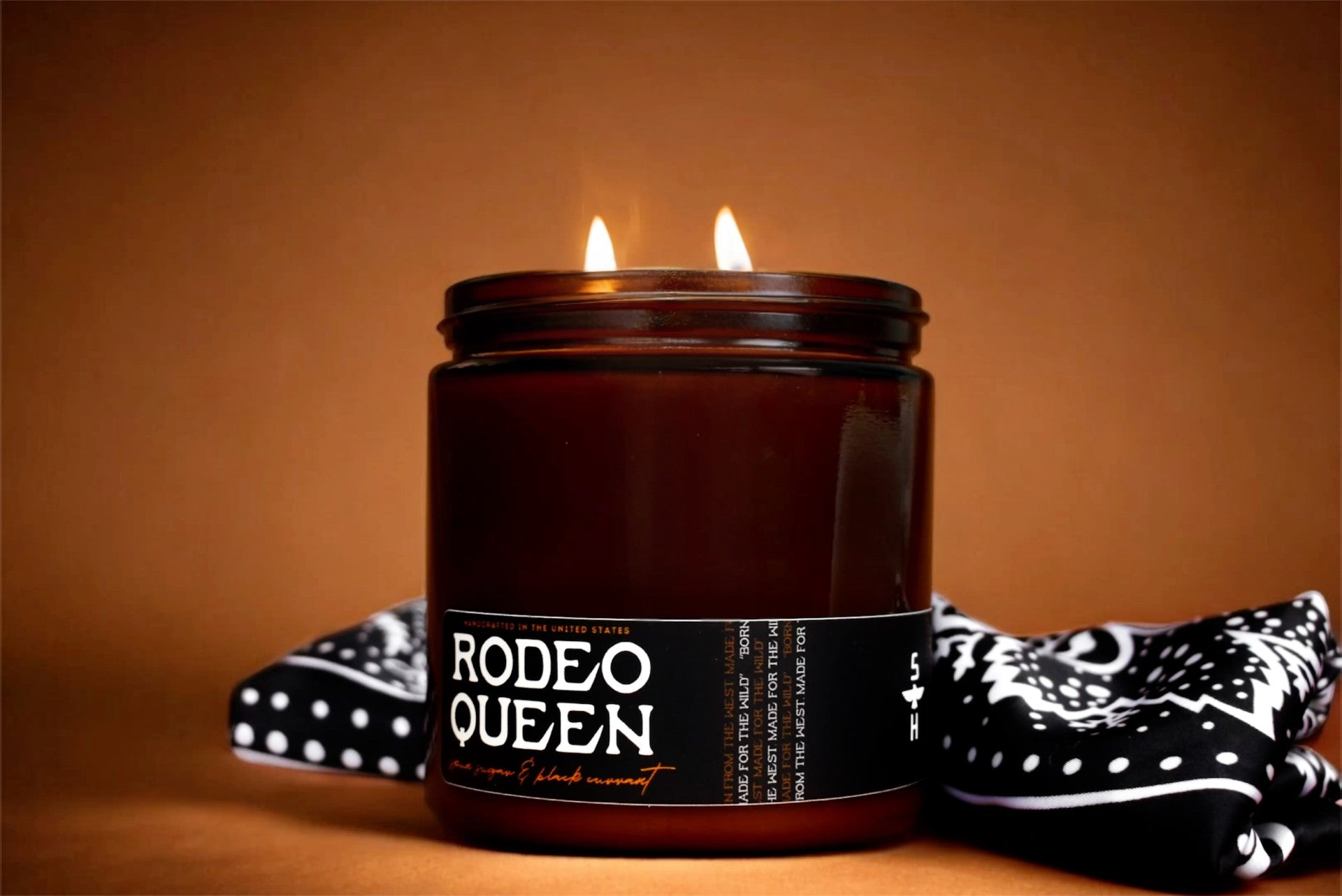 A lit brown glass candle labeled "Black Currant & Spun Sugar" by Faire sits on a brown surface, with a black and white patterned bandana draped behind it. The background features a warm brown shade.