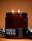A lit brown glass candle labeled "Black Currant & Spun Sugar" by Faire sits on a brown surface, with a black and white patterned bandana draped behind it. The background features a warm brown shade.