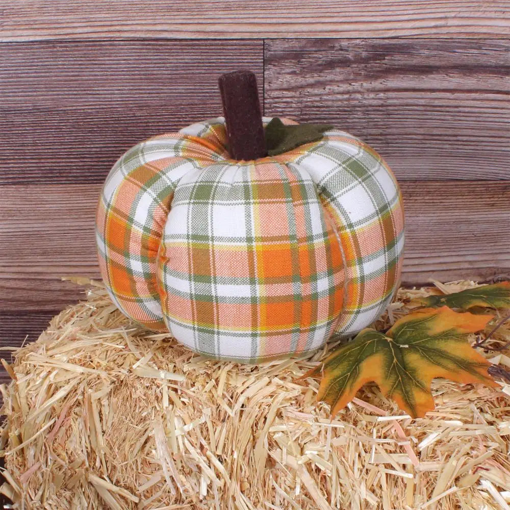 The Faire Pumpkin is a fabric plaid fall decor piece, ideal as a farmhouse ornament. It sits on a hay bale with two artificial autumn leaves, set against a wooden plank background.