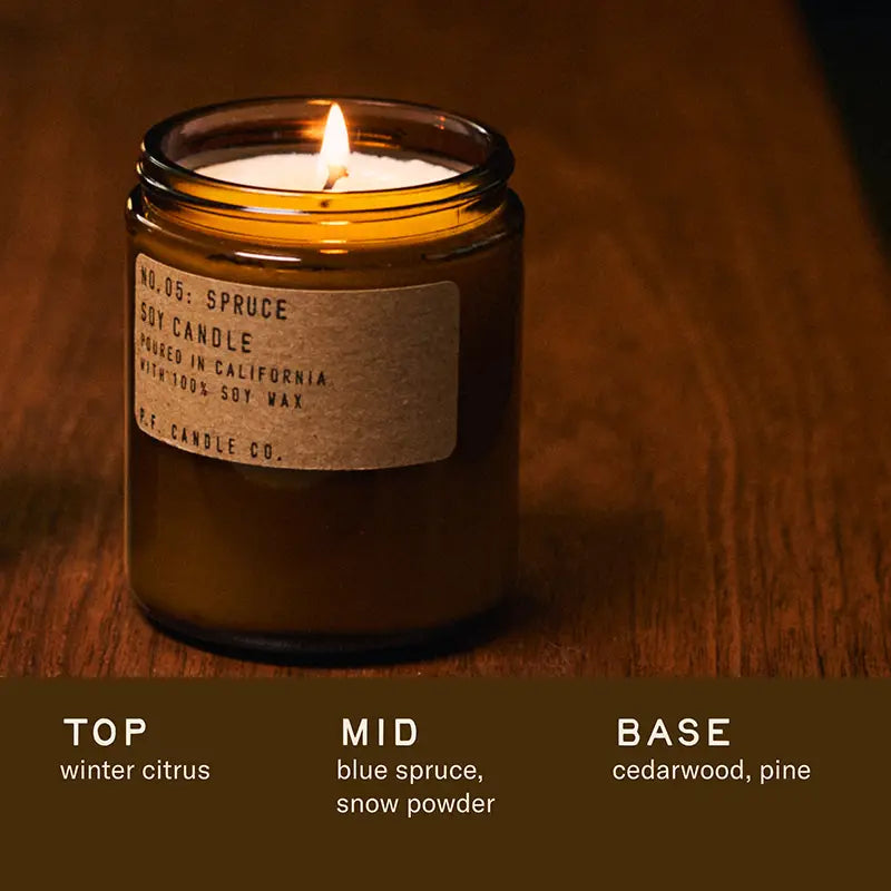 A lit PFC Candle by Faire, in a brown glass jar labeled &quot;No. 05: Spruce,&quot; sits on wood. Made with soy wax and fine fragrance oils, it features notes of winter citrus, blue spruce, snow powder, cedarwood, and pine.