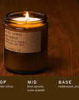 A lit PFC Candle by Faire, in a brown glass jar labeled "No. 05: Spruce," sits on wood. Made with soy wax and fine fragrance oils, it features notes of winter citrus, blue spruce, snow powder, cedarwood, and pine.